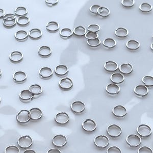 Puede incluir: Primer plano de pequeños anillos de salto plateados dispersos sobre una superficie blanca. Estos anillos abiertos se utilizan para la fabricación de joyas u otras manualidades. Cada anillo mide aproximadamente 0,5 cm de diámetro.