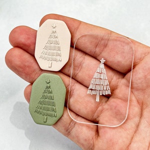 Op de afbeelding: Twee kerstboomvormige stempels van polymeerklei, een in groen en een in beige, met een transparante acryl stempel op de achtergrond. De stempels hebben een ster bovenop en een textuurontwerp.