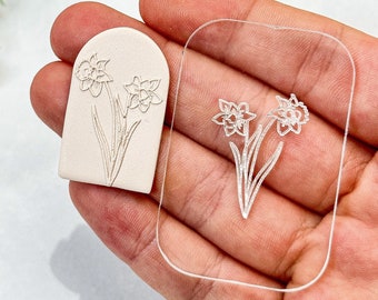 Puede incluir: Un sello de cerámica blanca con un diseño en relieve de narcisos. El sello se sostiene en una mano junto a un sello de acrílico transparente con el mismo diseño.