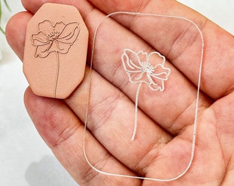 Puede incluir: Un sello de acrílico transparente con un dibujo lineal de una flor. El sello se sostiene en una mano. El sello se utiliza para manualidades y scrapbooking.