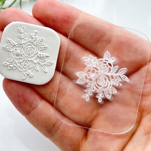 Könnte beinhalten: Ein weißer Stempel mit einem filigranen Blumenmuster und einem transparenten Acrylblock. Der Stempel besteht aus einem weichen, biegsamen Material.