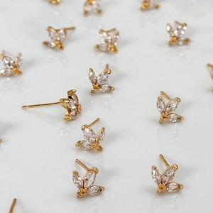 Könnte beinhalten: Eine Sammlung von goldenen Ohrsteckern, die jeweils aus drei in einer Gruppe angeordneten, klar geschliffenen Marquise-Steinen bestehen. Die Ohrstecker haben einen Stiftverschluss und sind in einem Raster angeordnet, das die goldene Fassung zeigt.