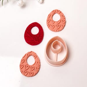 May include: A collection of polymer clay earring components and a cutter. Two peach-colored oval earrings with heart patterns, a red textured oval earring, and a peach-colored cutter with an oval shape.
