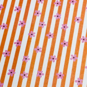 Peut inclure: Une feuille de papier rectangulaire présentant un motif répétitif de rayures verticales orange et blanches. De petites fleurs roses à cinq pétales avec des centres sombres sont réparties uniformément sur la surface, recouvrant les rayures.