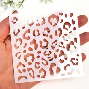 Puede incluir: Un esténcil cuadrado blanco con un diseño de estampado de leopardo. El esténcil se sostiene en una mano, con el diseño recortado. El fondo es blanco, con ramitas de flores blancas.