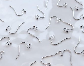 May include: A collection of silver-colored earring hooks with small ball accents, scattered on a white, slightly reflective surface. The hooks are made of thin, curved metal wire, designed for jewelry making and crafting.