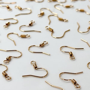 May include: A collection of gold-colored earring hooks, arranged on a white surface. The hooks have a curved design with a small coil and a loop at the end. The hooks are made of a shiny, metallic material.