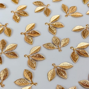 May include: A collection of small, gold-toned metal charms, each shaped like a cluster of three textured leaves. These individual leaf charms feature intricate vein detailing on their surfaces and include a small loop at the base for attachment.