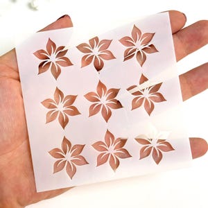 Peut inclure: Une feuille de pochoir blanche avec neuf motifs de fleurs. Chaque fleur a six pétales et une couleur marron clair. Le pochoir est tenu dans une main sur un fond blanc, avec des fleurs blanches en arrière-plan.