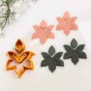 Peut inclure: Collection de boucles d'oreilles florales faites à la main et un emporte-pièce en forme de fleur. Les boucles d'oreilles sont corail et vert foncé, avec des textures de feuilles. L'emporte-pièce est orange.