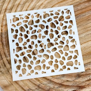Op de afbeelding: Een vierkant wit sjabloon met een herhalend patroon van onregelmatige, afgeronde vormen die doen denken aan een giraffepatroon of verspreide kiezels. Het sjabloon is dun, plat en flexibel, met uitsparingen die de negatieve ruimte vormen.