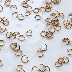 Op de afbeelding: Een verzameling kleine, goudkleurige open springringen verspreid op een wit oppervlak. De ringen zijn in verschillende richtingen georiënteerd, sommige open en andere gesloten, voor het maken van sieraden of knutselen.