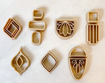 May include: Set of eight cookie cutters in a beige color, featuring various geometric and abstract shapes, including a diamond, a flower, and a teardrop.