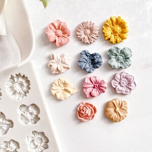 May include: Assortment of 10 colourful flower-shaped resin embellishments in various colours, including pink, yellow, blue, and green. The embellishments are arranged on a white surface.
