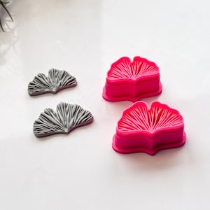 May include: Two gray ginkgo leaf-shaped molds and two bright pink ginkgo leaf-shaped cookie cutters. The molds have detailed vein patterns, while the cutters have a simpler design. The items are arranged on a white surface.