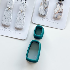 Peut inclure: Deux paires de boucles d'oreilles en argile polymère faites à la main. Une paire est blanche avec des points noirs et l'autre paire est blanche avec un marbrage gris et or. Les boucles d'oreilles sont présentées sur un fond blanc avec une carte de visite qui dit "Tera Clay Handmade Earrings" et une adresse de site Web. Deux moules en silicone turquoise sont également présentés sur l'image.