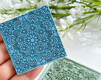 May include: Two square, floral-patterned stamps in shades of blue and green. The stamps feature a detailed mandala design with flowers and geometric shapes. The blue stamp is held in a hand, while the green stamp rests on a white surface.