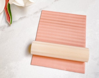 May include: A pink and white silicone rolling pin with a chevron pattern. The rolling pin is rolled up on a flat sheet of pink silicone with the same chevron pattern.