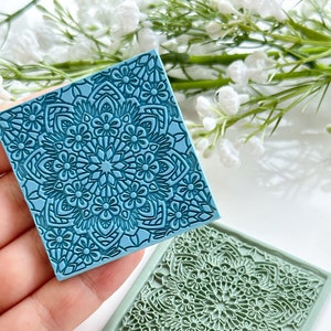 Op de afbeelding: Twee vierkante stempels met bloempatronen in blauw en groen. De stempels hebben een gedetailleerd mandala-ontwerp met bloemen en geometrische vormen. De blauwe stempel wordt in een hand gehouden, terwijl de groene stempel op een wit oppervlak ligt.