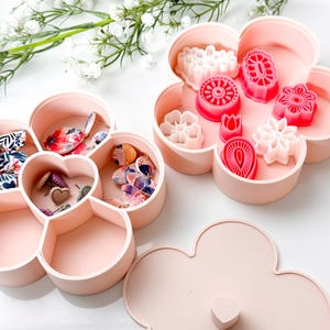 Puede incluir: Contenedores de almacenamiento rosas en forma de flor con compartimentos, llenos de suministros de artesanía y cortadores de galletas. Los contenedores están abiertos, revelando el contenido, y una tapa a juego está cerca. Flores blancas en el fondo.