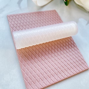 Puede incluir: Un rodillo de silicona blanco con un patrón de diamante en una estera de silicona rosa con un patrón de diamante.