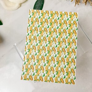 May include: A white card with a repeating pattern of yellow corn on the cob with green husks. The corn is arranged in rows and is set against a white background.