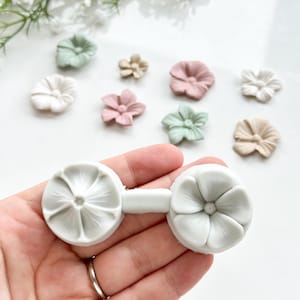 Può includere: Uno stampo in silicone bianco per realizzare forme di fiori bidimensionali. Lo stampo ha due forme di fiori collegate da una striscia sottile. Lo stampo è tenuto in una mano.