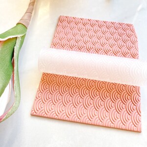 May include: A pink silicone rolling pin with a white silicone rolling mat. The mat has a repeating pattern of overlapping half circles.