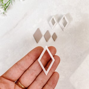 May include: Set of five white plastic diamond-shaped cookie cutters. The cutters are of varying sizes and are arranged on a white surface. The largest cutter is being held in a hand.