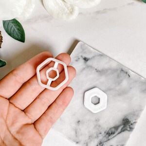 May include: Two white plastic cookie cutters in the shape of a hexagon with a smaller hexagon inside. The larger cutter is being held in a hand, and the smaller cutter is on a marble surface.