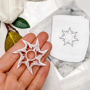 Op de afbeelding: Witte plastic koekjesvormer in de vorm van een zon met een ring in het midden. De vormer wordt in een hand gehouden. Een tweede, grotere zonvormige koekjesvormer is zichtbaar op de achtergrond.