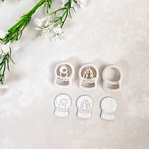 Op de afbeelding: Zes witte plastic koekjesvormen in de vorm van sneeuwbollen. De sneeuwbollen hebben verschillende ontwerpen, waaronder een sneeuwpop, een dennenboom en een leeg ontwerp.