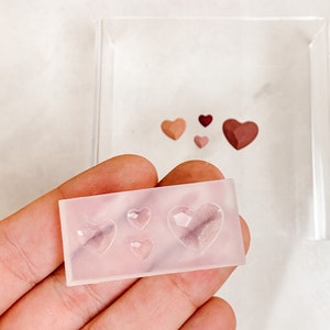May include: A clear silicone mold with five heart-shaped cavities. The mold is being held in a hand. There are four heart-shaped resin pieces in the background, in various shades of pink and brown.