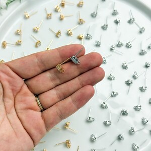 May include: A collection of gold and silver-toned earring posts with small loops for attaching charms. The earring findings feature a prong setting for a single stone and a thin metal post with a loop at the base, arranged on a white surface.