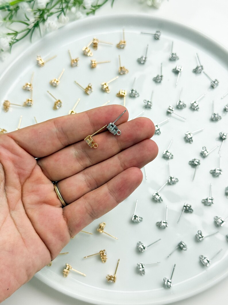 May include: A collection of gold and silver-toned earring posts with small loops for attaching charms. The earring findings feature a prong setting for a single stone and a thin metal post with a loop at the base, arranged on a white surface.