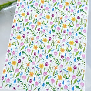 Pode incluir: Um papel branco com um padrão floral em aquarela. O design apresenta narcisos amarelos, tulipas roxas, tulipas rosas, muscaris azuis e folhas verdes. As flores estão dispostas em um padrão repetitivo.