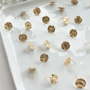 May include: A collection of 20 gold-toned metal earring blanks with a textured surface. Each blank has a small hole in the center and a post on the back.
