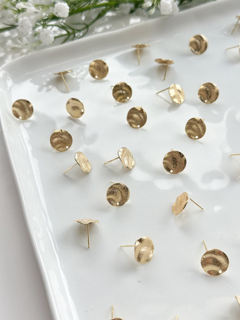 May include: A collection of 20 gold-toned metal earring blanks with a textured surface. Each blank has a small hole in the center and a post on the back.