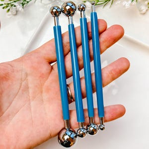 Puede incluir: Juego de cuatro herramientas con puntas de bola azules y plateadas. Cada herramienta tiene una bola plateada brillante en un extremo y una bola o punta plateada más pequeña en el otro, conectadas por un mango azul. Herramientas para manualidades o escultura.