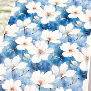 May include: A floral print featuring white flowers with brown centers against a blue watercolor background. The flowers are detailed with delicate petals and are arranged in a repeating pattern. The overall aesthetic is soft and artistic.