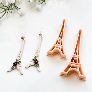 以下が含まれることがあります： 赤い花があしらわれた白いエッフェル塔のイヤリングのペア。イヤリングの隣には、ピンクのプラスチック製のエッフェル塔のクッキーカッターが2つあります。