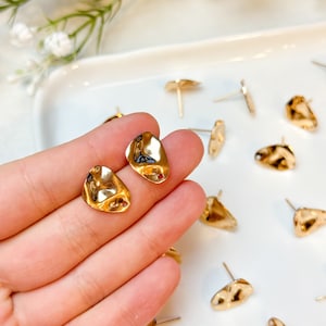 May include: Gold-colored stud earrings with an irregular, textured surface. The earrings have a post backing and are displayed on a white surface. The earrings are small and appear to be made of metal.