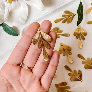 Peut inclure: Gros plan sur une main tenant un pendentif en forme de feuille dorée. Le pendentif est en métal et a une surface texturée. Le pendentif est entouré d'autres pendentifs similaires sur une surface blanche.