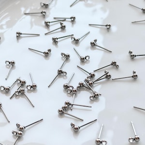 May include: A collection of silver-colored stud earrings with a three-ball design, scattered on a white surface. Each earring has a post for insertion and a cluster of three small, round beads. Some earrings have clear, sparkling accents.
