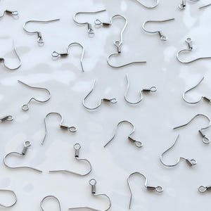Könnte beinhalten: Eine Ansammlung silberfarbener Ohrhaken, verstreut auf einer weißen Oberfläche. Jeder Haken hat ein gebogenes Design mit einer kleinen Feder und einer Schlaufe an der Basis, ideal für die Schmuckherstellung. Die Haken bestehen aus glänzendem Metall.