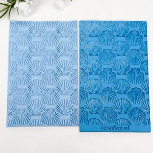 Può includere: Due tappetini rettangolari in silicone con texture a motivo ripetuto di conchiglie. Il tappetino a sinistra è azzurro, quello a destra è blu più scuro con il testo teraclay.nl stampato in piccole lettere sul bordo inferiore della superficie.