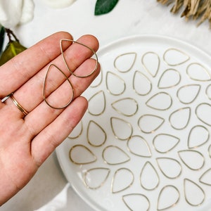 Op de afbeelding: Goudkleurige, druppelvormige sieradencomponenten. Twee worden in een hand gehouden, met vele anderen gerangschikt op een wit bord. De druppels zijn delicaat en hebben een eenvoudig, elegant ontwerp, geschikt voor het maken van oorbellen of hangers.