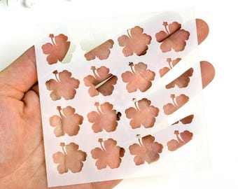 May include: A clear plastic stencil featuring a repeating pattern of hibiscus flower cutouts. The stencil is held in a hand against a white background, with a few sprigs of white flowers in the upper left corner. The hibiscus flowers are a light brown color.