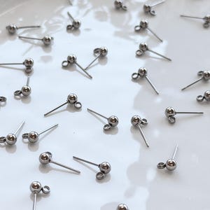 Peut inclure: Plusieurs boucles d'oreilles clous argentées, chacune avec une boule et une tige pour percer l'oreille. Elles sont disposées sur une surface blanche. Les boucles d'oreilles sont probablement en acier inoxydable. Bijoux discrets et élégants.