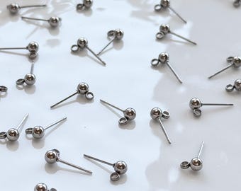 May include: A collection of small, silver-colored stud earrings with a ball-shaped design and a post for piercing. The earrings are scattered across a white surface, creating a minimalist aesthetic. The earrings are likely made of stainless steel.